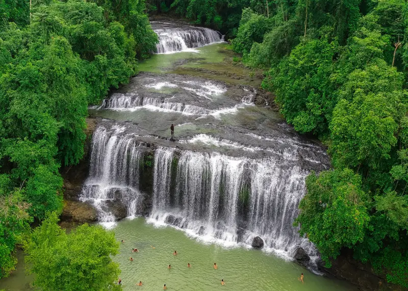 philippines multi tier waterfalls Multi-tier waterfall in the Philippine jungle with locals standing on rocks and lush greenery surrounding.